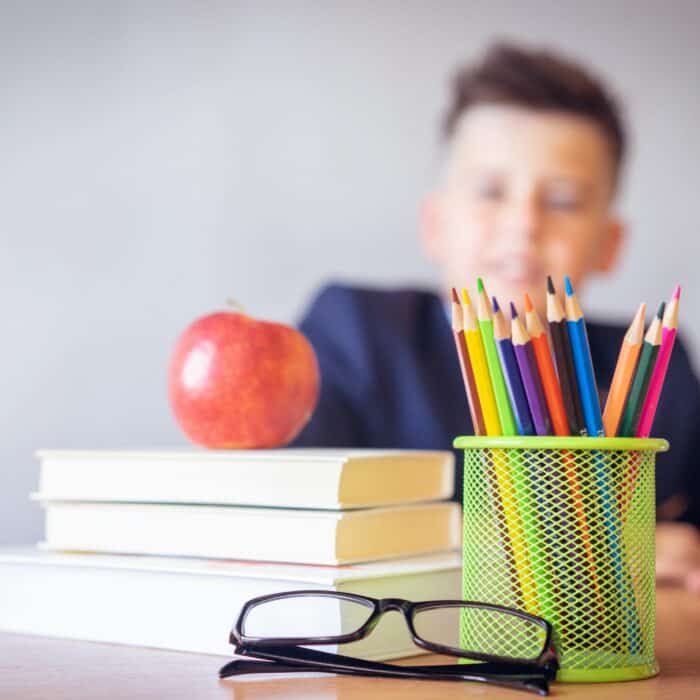 Schoolboy smiling behind a desk with books, pencils, and an alarm clock symbolizing study and creativity.